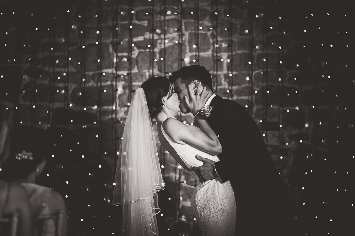 A couple kisses during their wedding photoshoot in front of a string of lights.