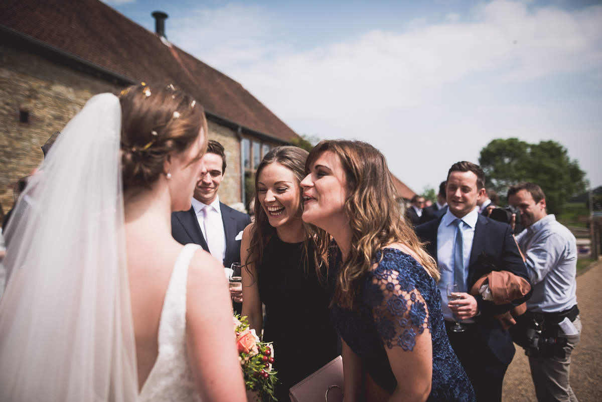 A bride, groom, and wedding photographer capturing a kiss between the bride and her bridesmaids in front of a barn.