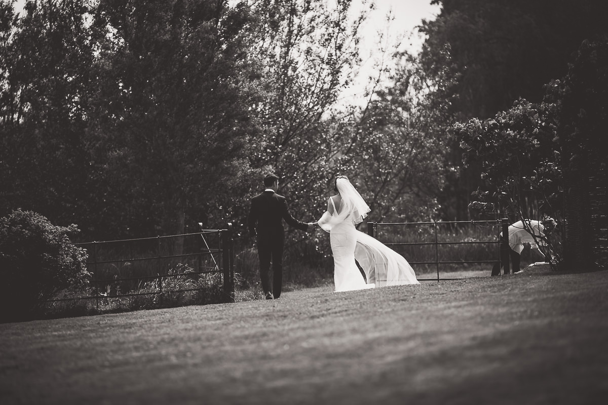 Wedding photographer captures black and white wedding photos of a bride and groom walking in the grass.