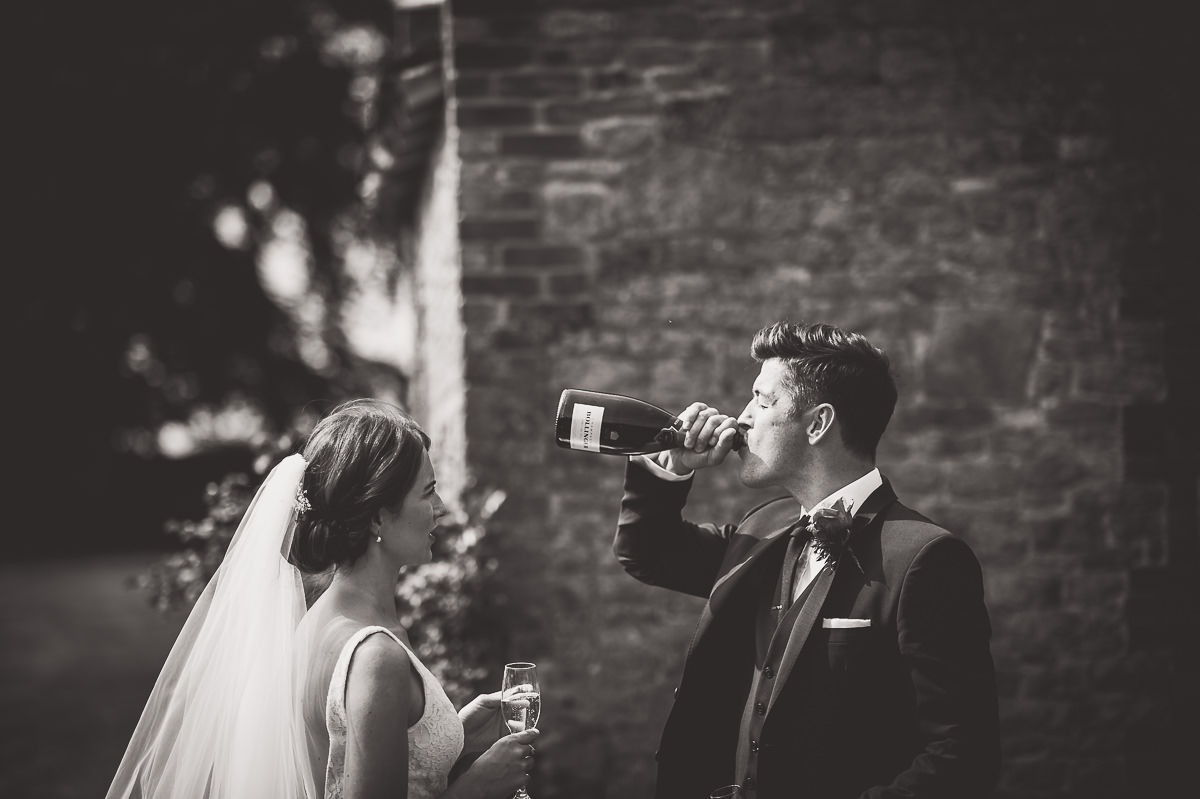 A wedding photographer captures the groom and bride sharing champagne in front of a brick wall for their wedding photos.