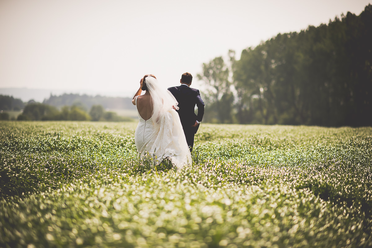 A wedding photographer captures a bride and groom strolling through a picturesque field.