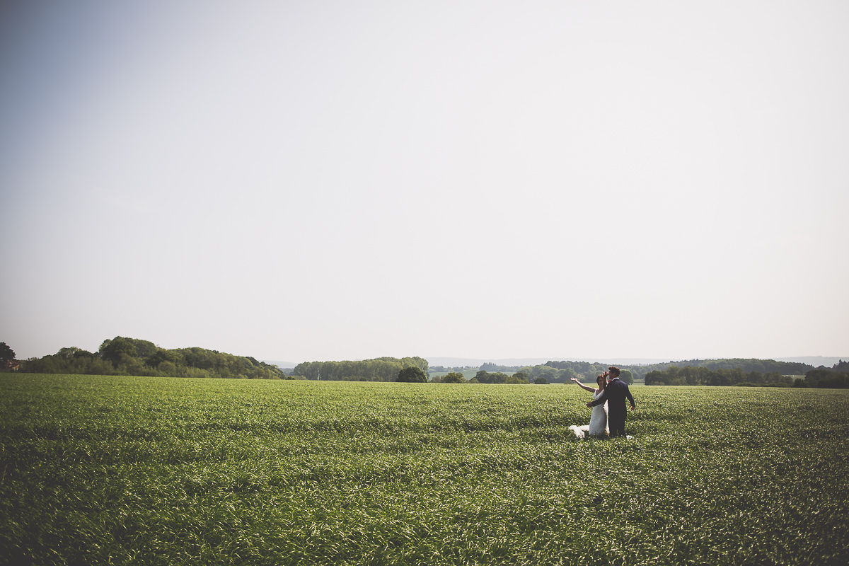 A wedding photographer captures a bride and groom standing in a field.