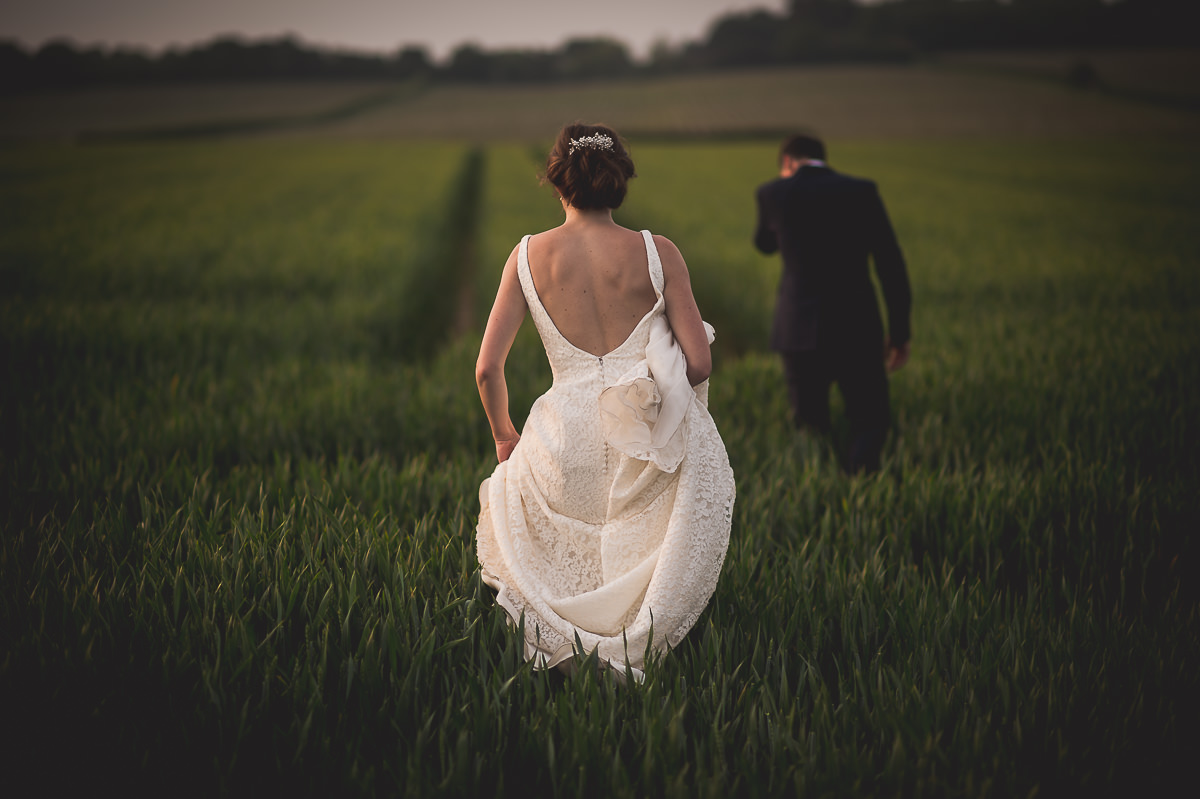 A wedding photographer captures a bride and groom strolling through a field.