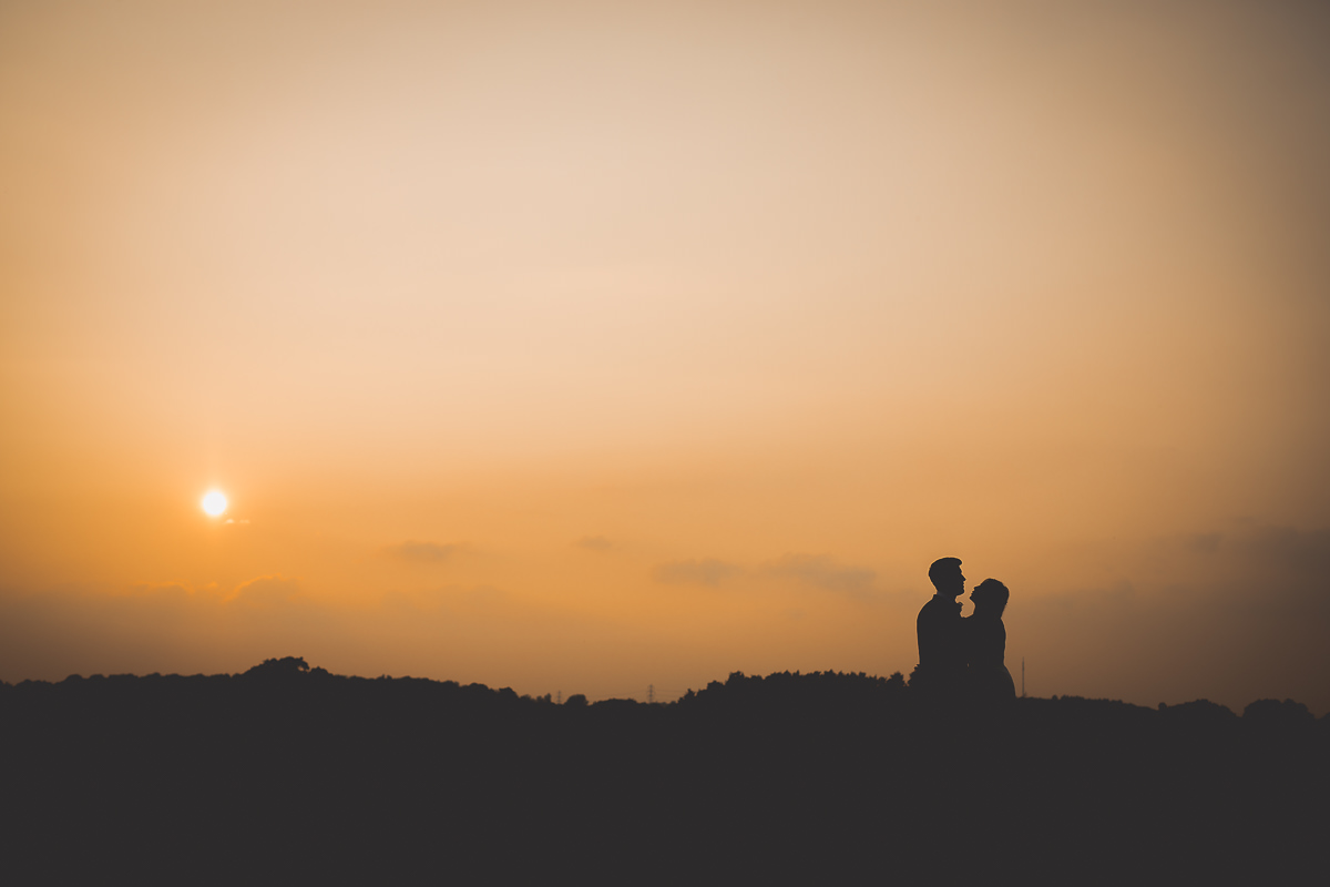 A groom, photographed by a wedding photographer, standing with his bride in front of a sunset.