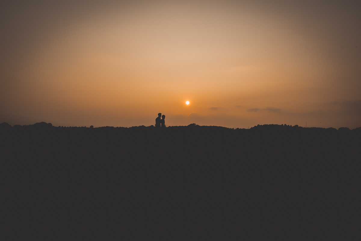 A groom and wedding photographer capture a breathtaking wedding photo with the couple standing on top of a hill at sunset.