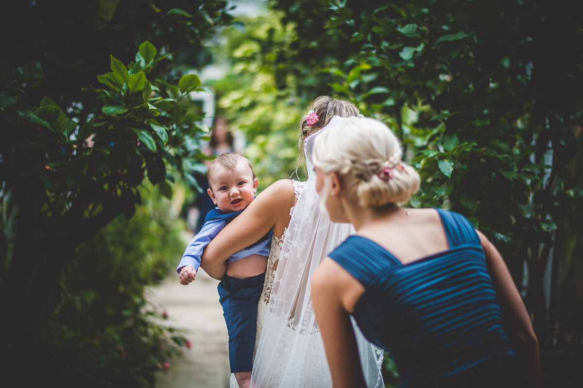 A bride, groom, and wedding photographer capture an endearing wedding photo as the bride walks down a path holding a baby.