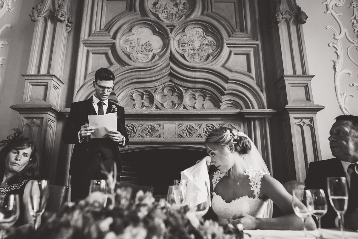 A wedding photographer captures a bride and groom reciting their vows in front of a lavish fireplace.