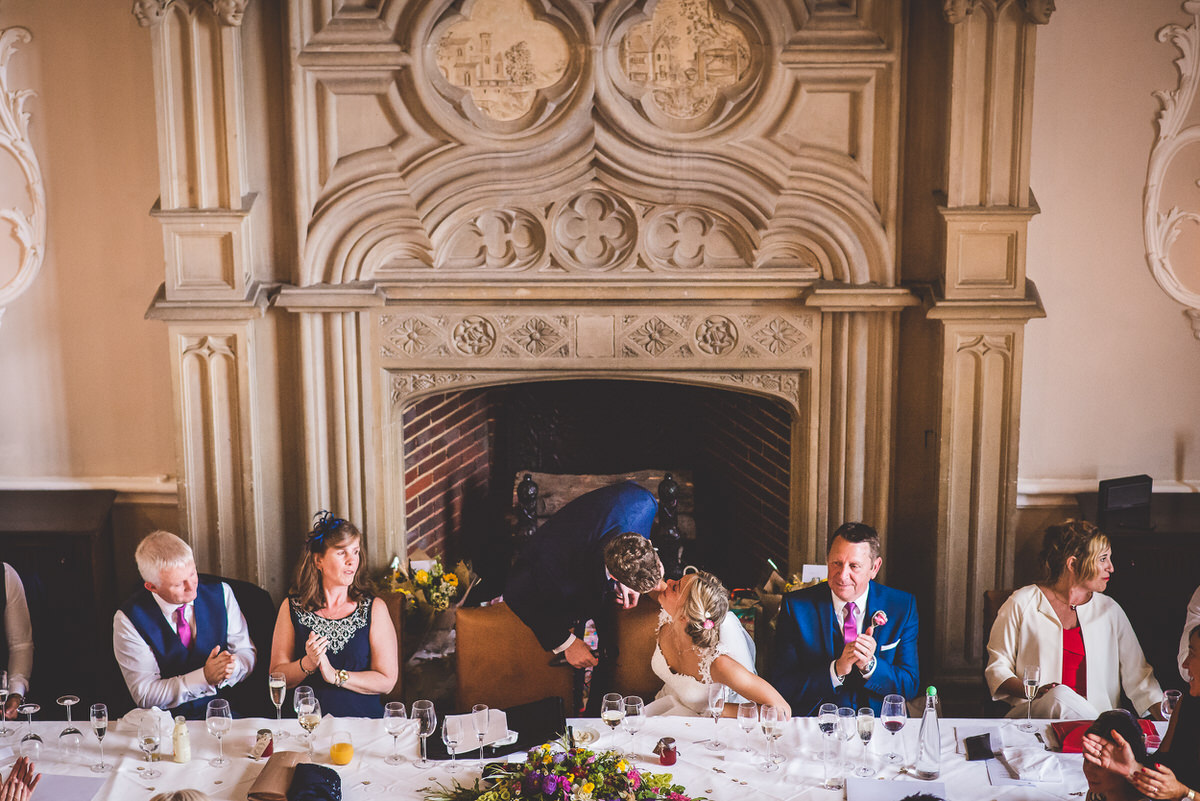 A wedding photographer capturing a group of people sitting at a table in front of a fireplace during a wedding.