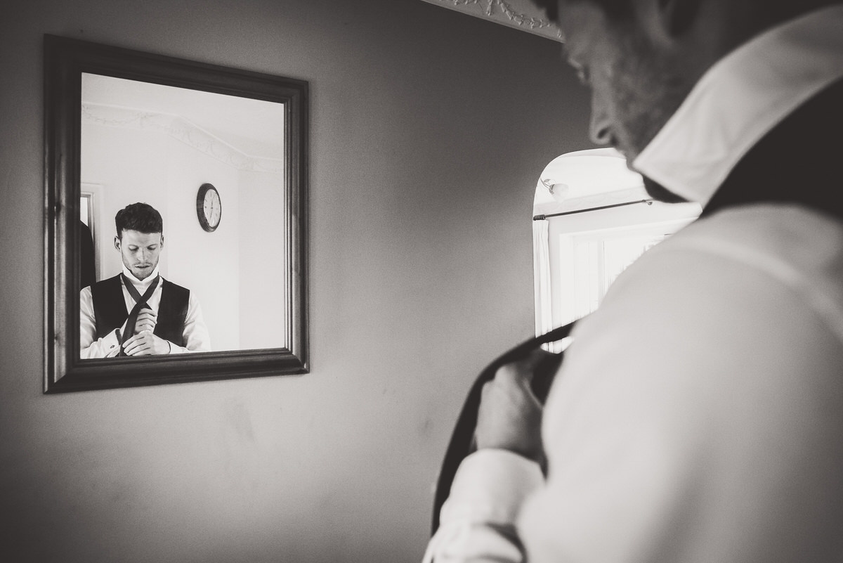 A man is preparing for a wedding, putting on his tie in front of a mirror.