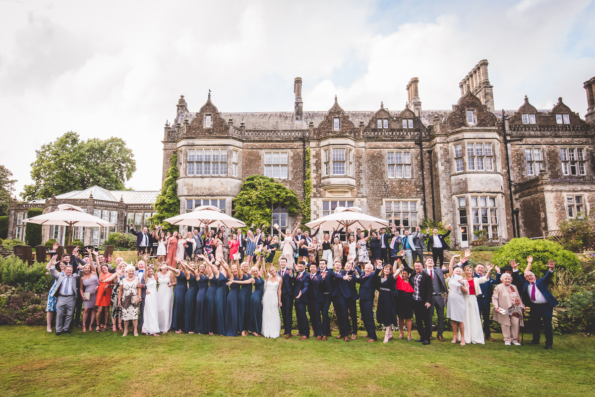 A bride and groom pose for their wedding photographer in front of a large mansion.