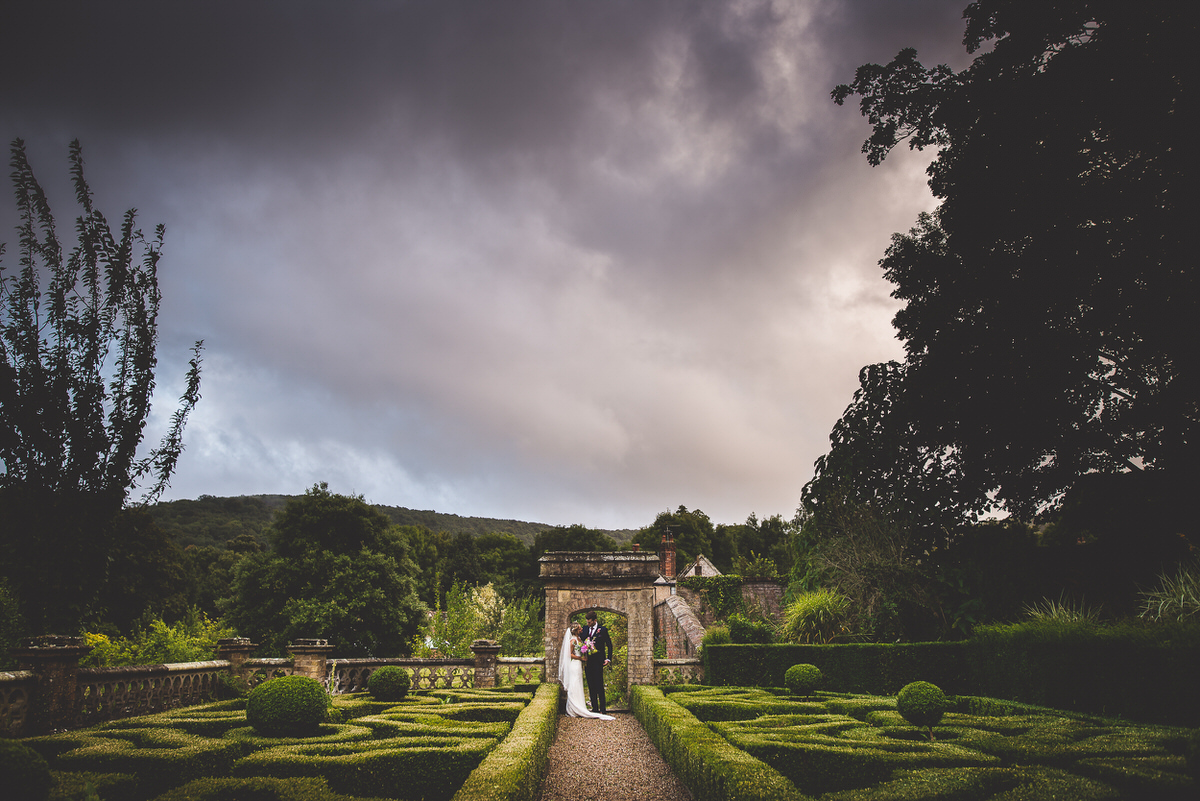 A wedding photographer captures a groom and bride in a garden beneath a stormy sky.