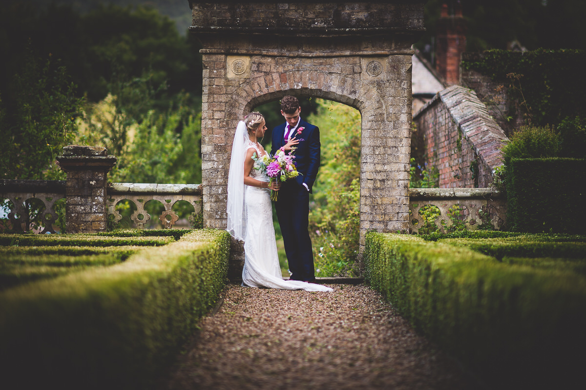 A wedding photo of a bride and groom standing in front of an archway in a garden.