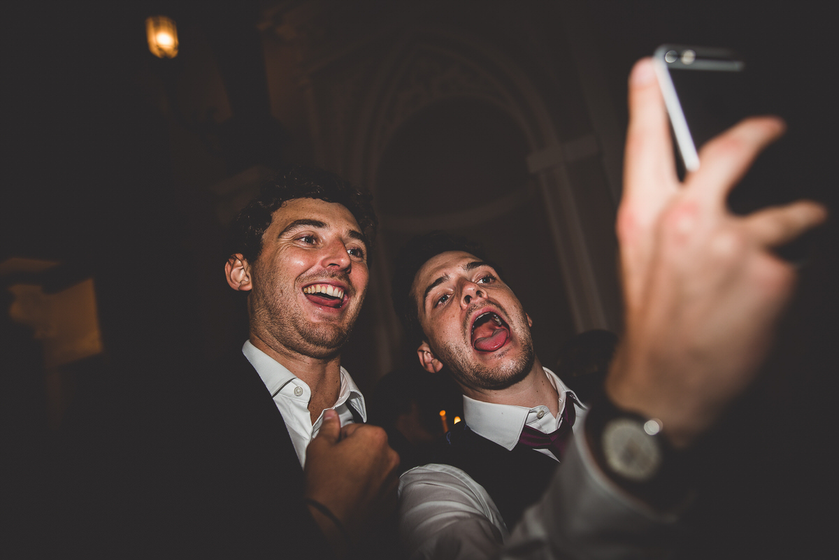 A groom and a wedding photographer capturing a selfie at a party.