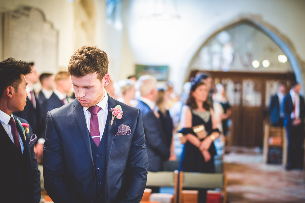 A groom, dressed in a suit, stands in a church while being photographed for his wedding album.