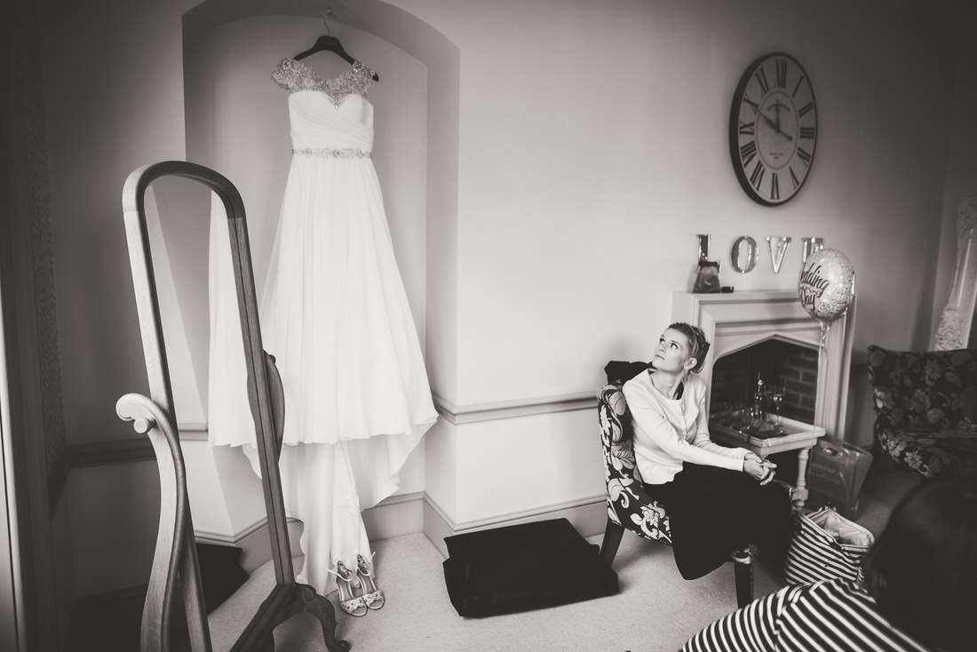 A wedding photographer captures a bride getting ready in a room with a dress hanging on the wall.