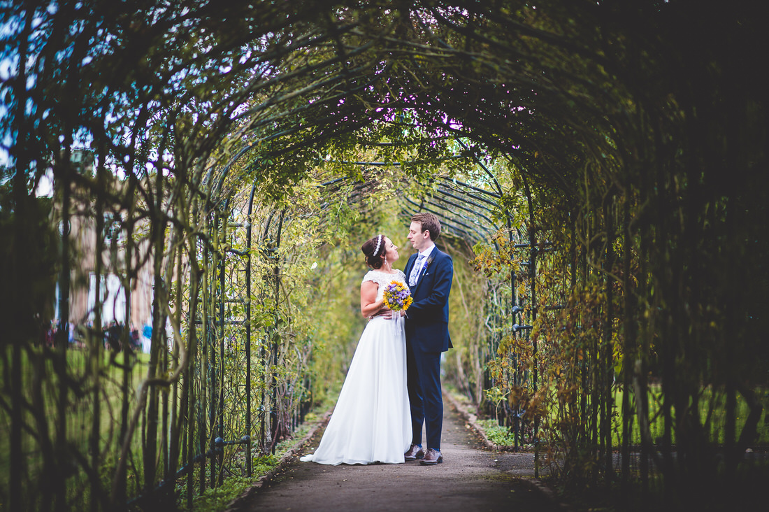 A wedding photographer captures a bride and groom posing for a photo in a garden archway.