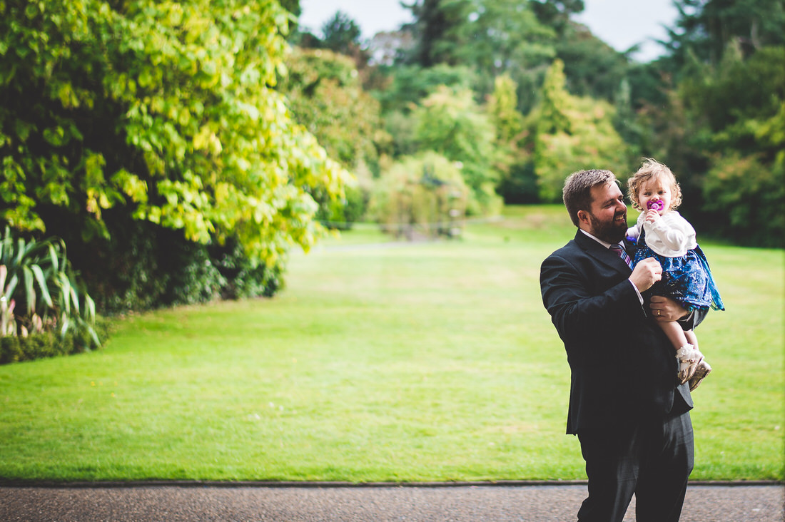 A wedding photographer capturing the groom holding a little girl in a park.