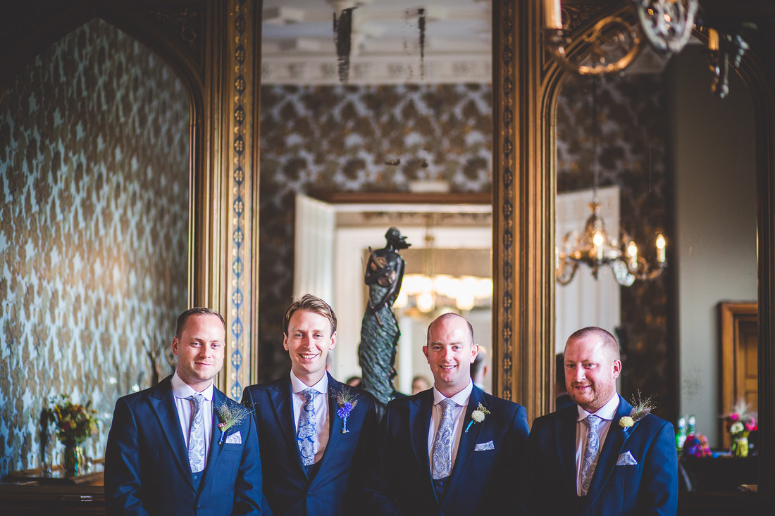 A group of groomsmen posing for a wedding photo in front of an ornate mirror.