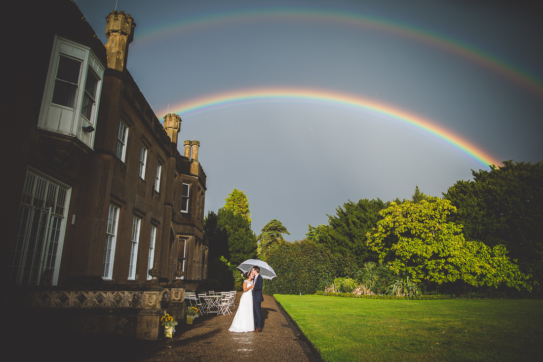 A bride and groom posing for their wedding photographer in front of a house with a rainbow backdrop.