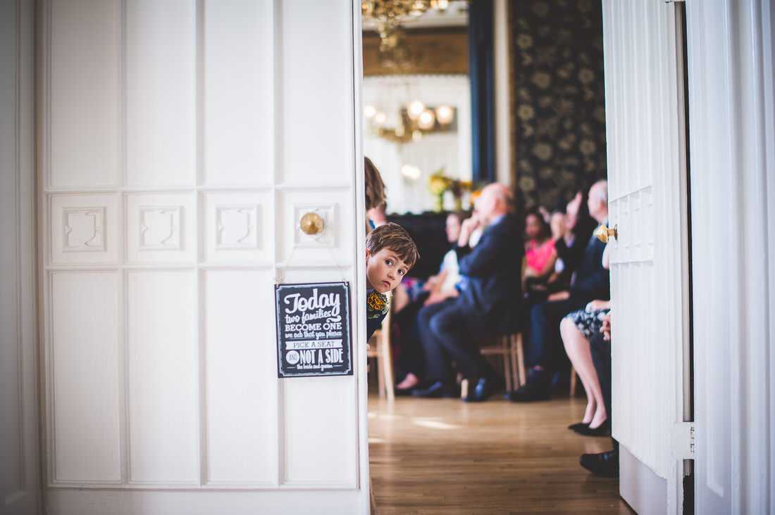 A little boy stands in a doorway, watching the bride and groom in a wedding ceremony.