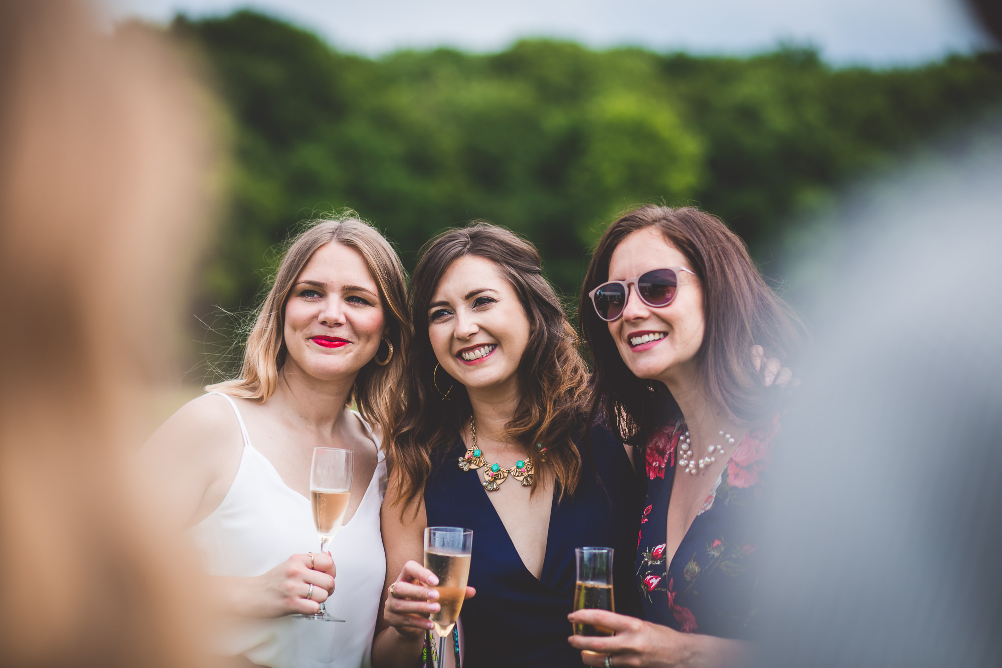 Three women, including the bride, holding champagne glasses in front of a group of people at a wedding celebration.