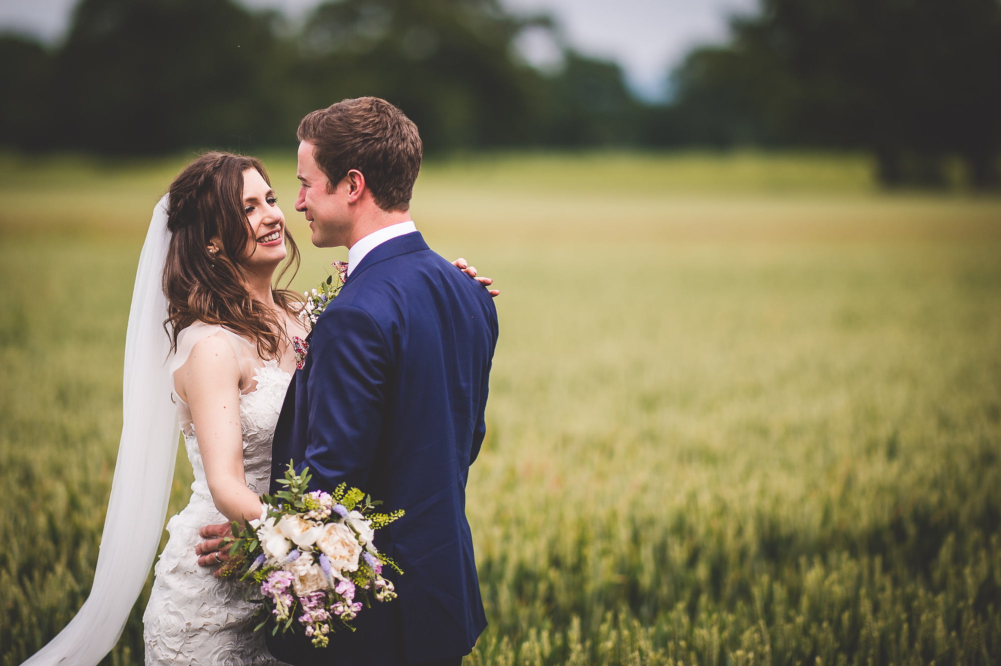 A wedding photographer captures the groom and bride standing in a wheat field.