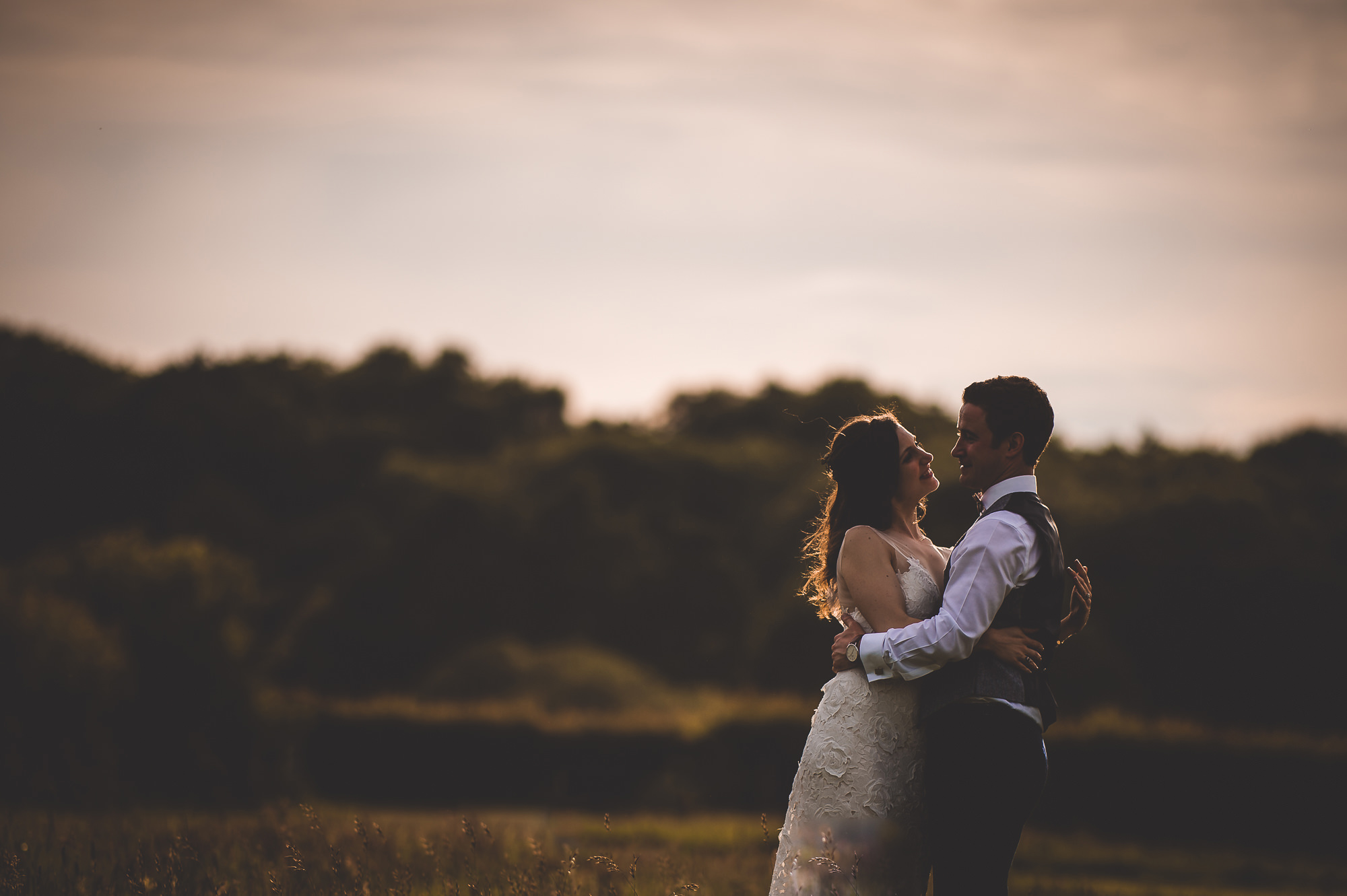 A wedding photographer captures a bride and groom embracing in a field at sunset.