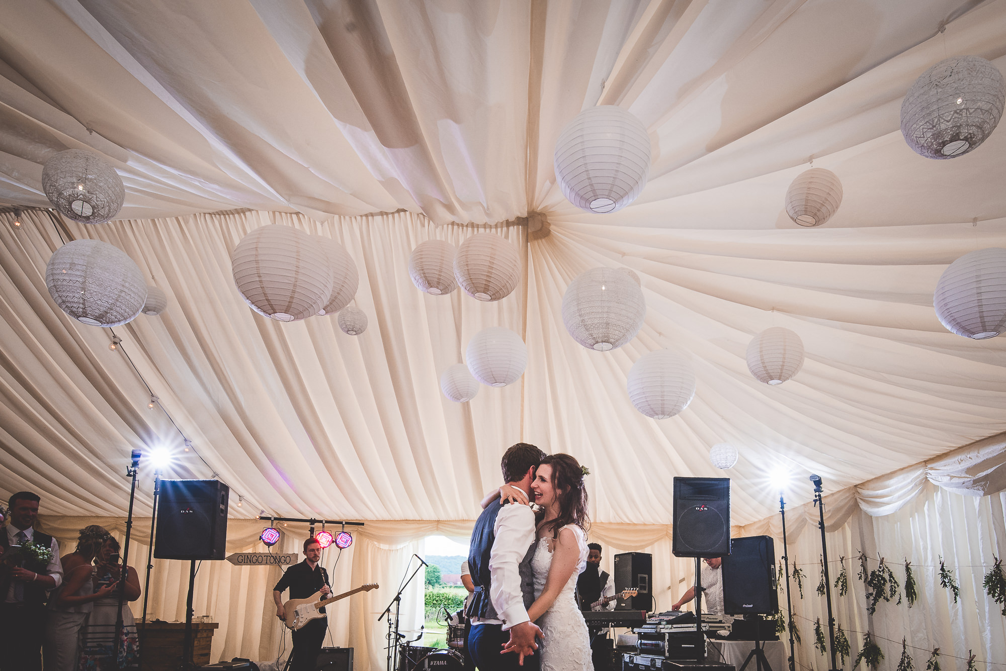 A wedding photographer captures a bride and groom sharing a kiss beneath paper lanterns in a tent.