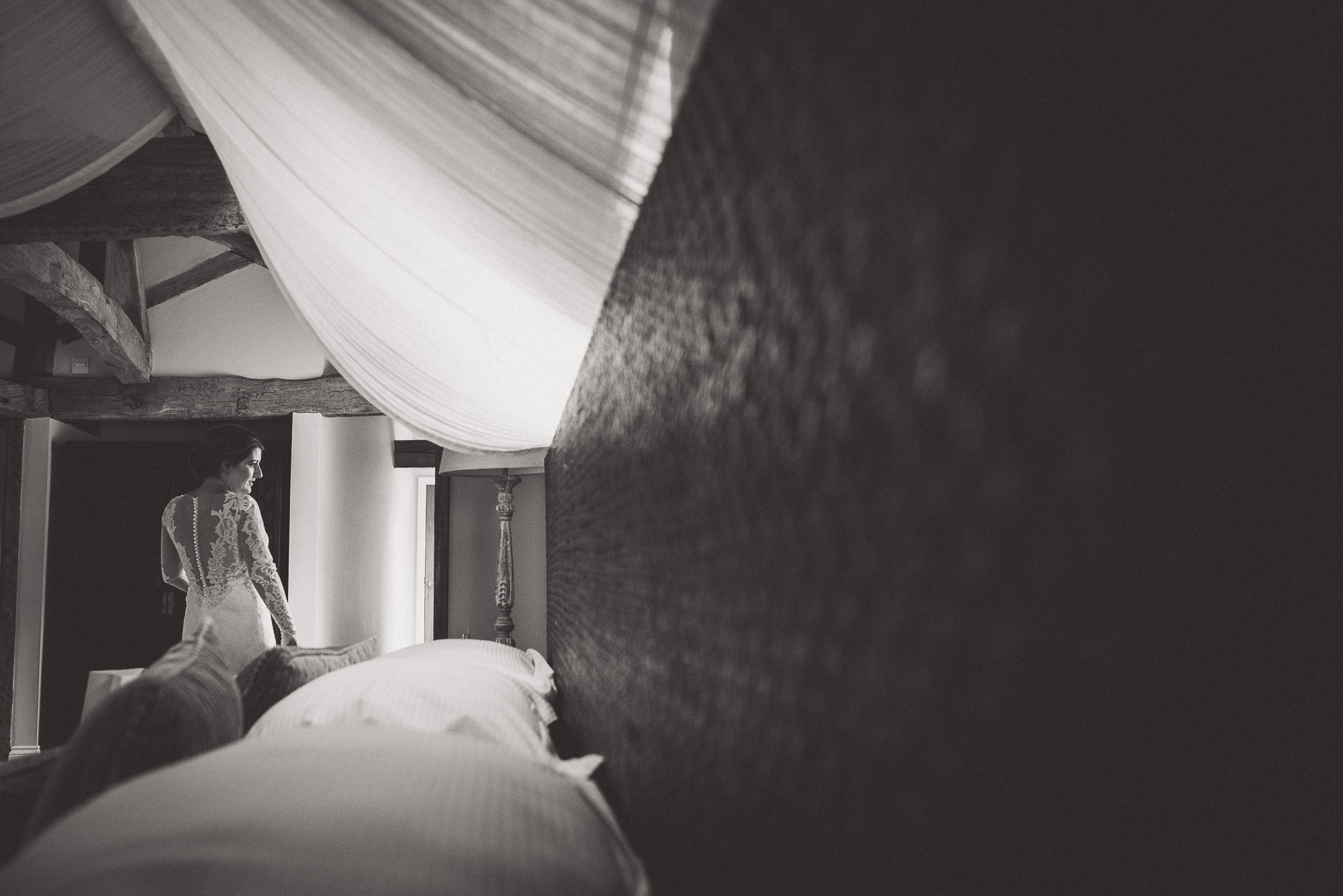 A bride and groom pose for their wedding photographer in a room with a bed.