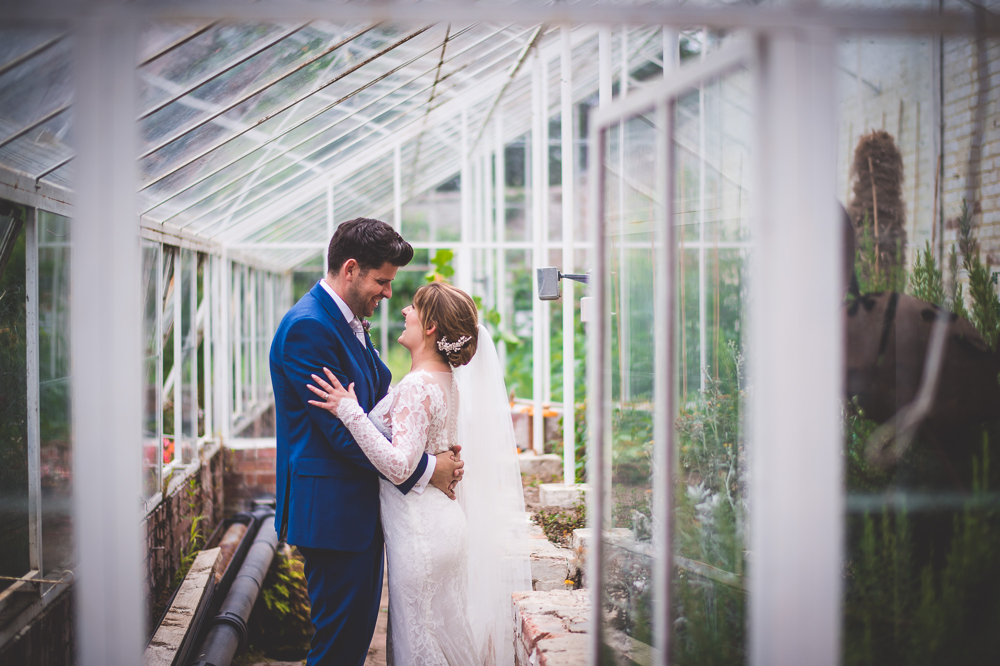 A wedding photographer capturing a groom embracing his bride in a greenhouse.