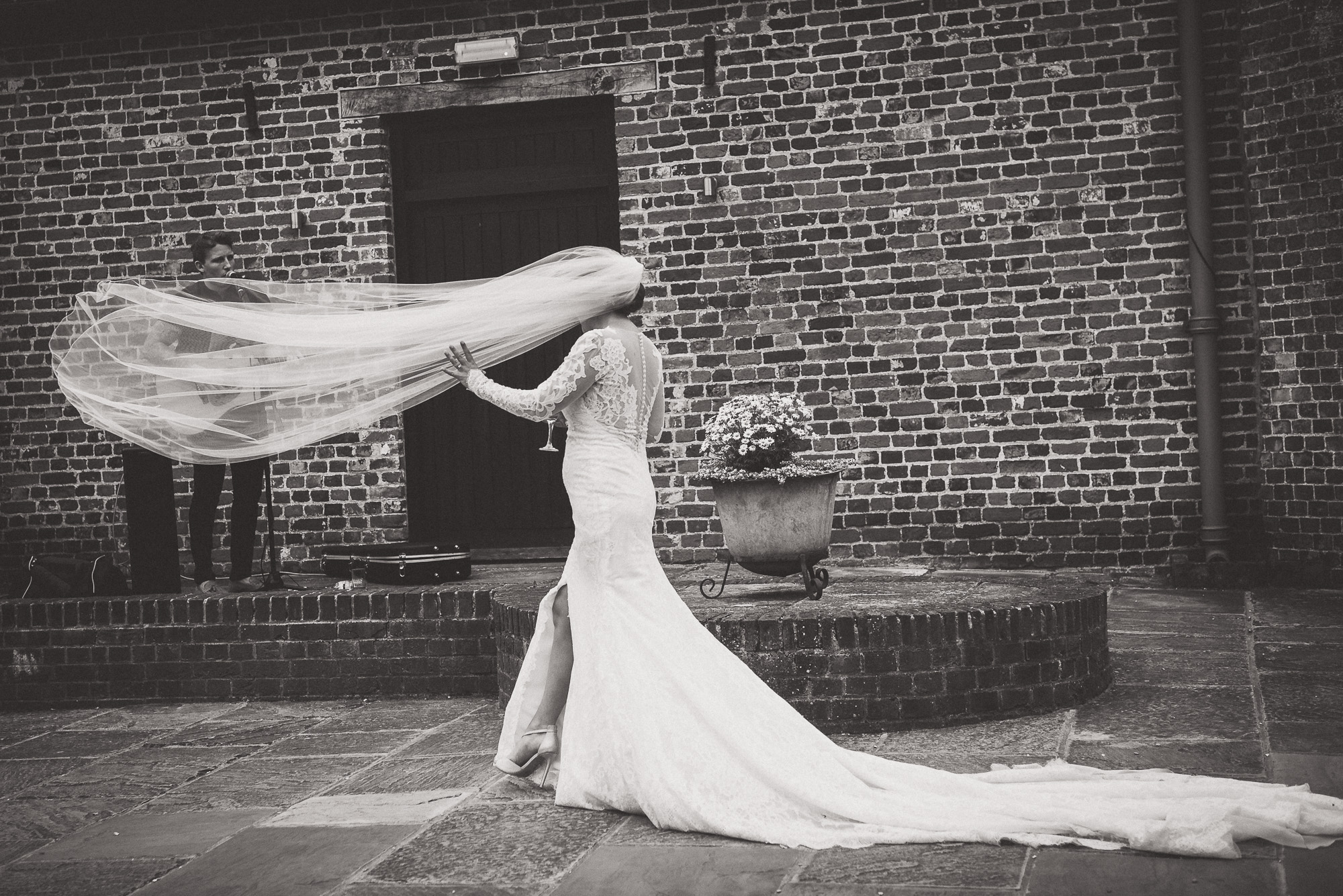 A bride poses for her wedding photo, blowing her veil in front of a brick building.