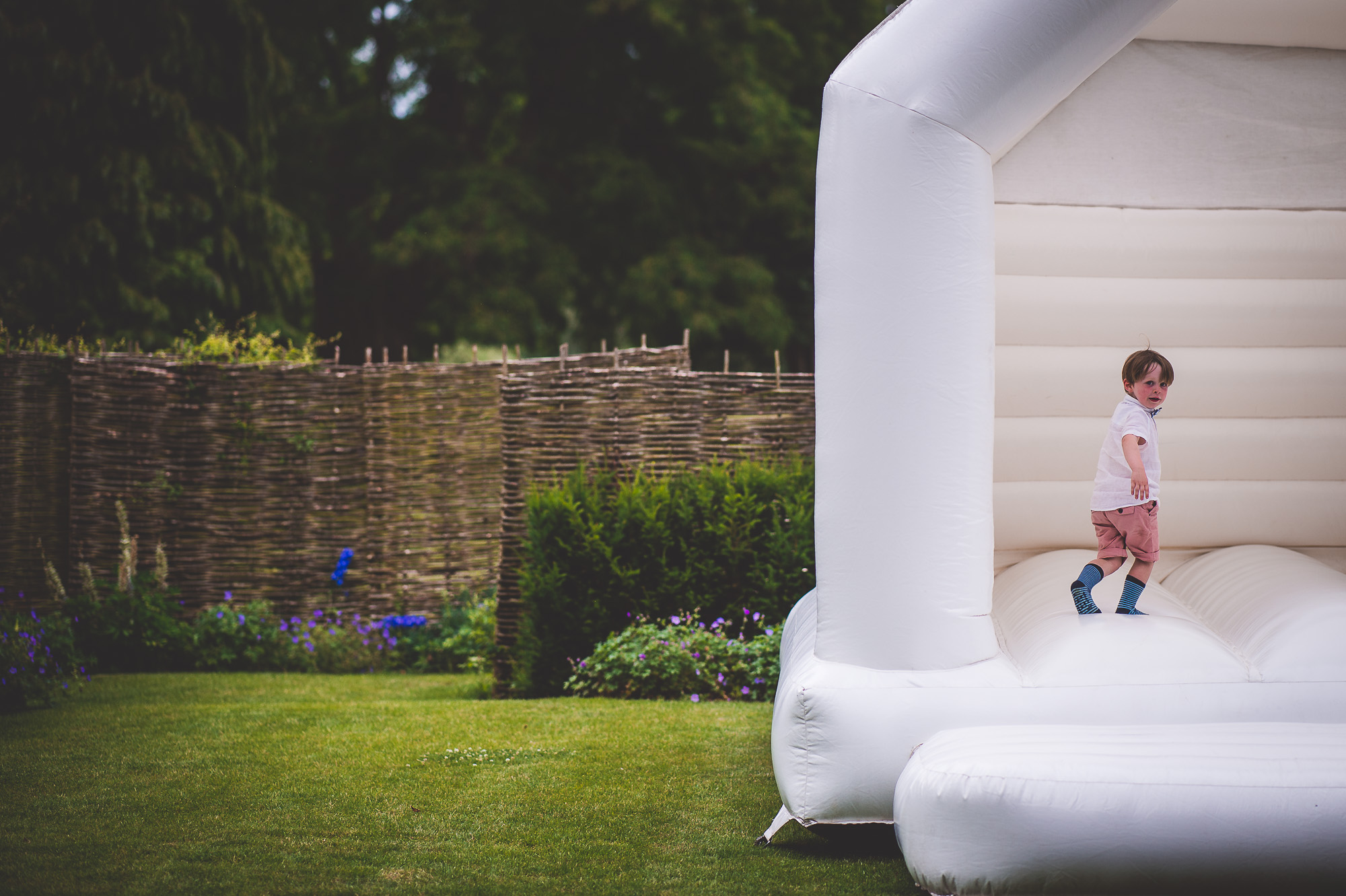 A child dressed as a groom poses on top of an inflatable bouncer in a garden for a wedding photographer's photo.