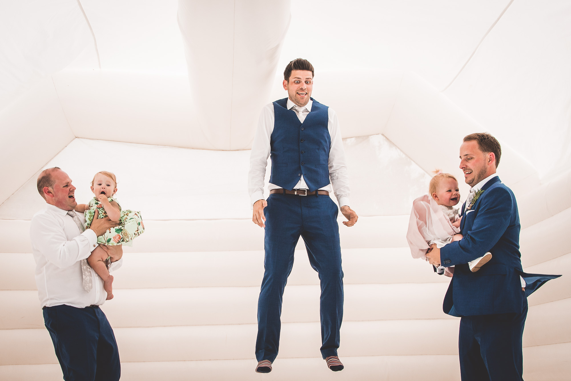 A group of groomsmen are jumping in an inflatable bouncy castle while the wedding photographer captures the moment.