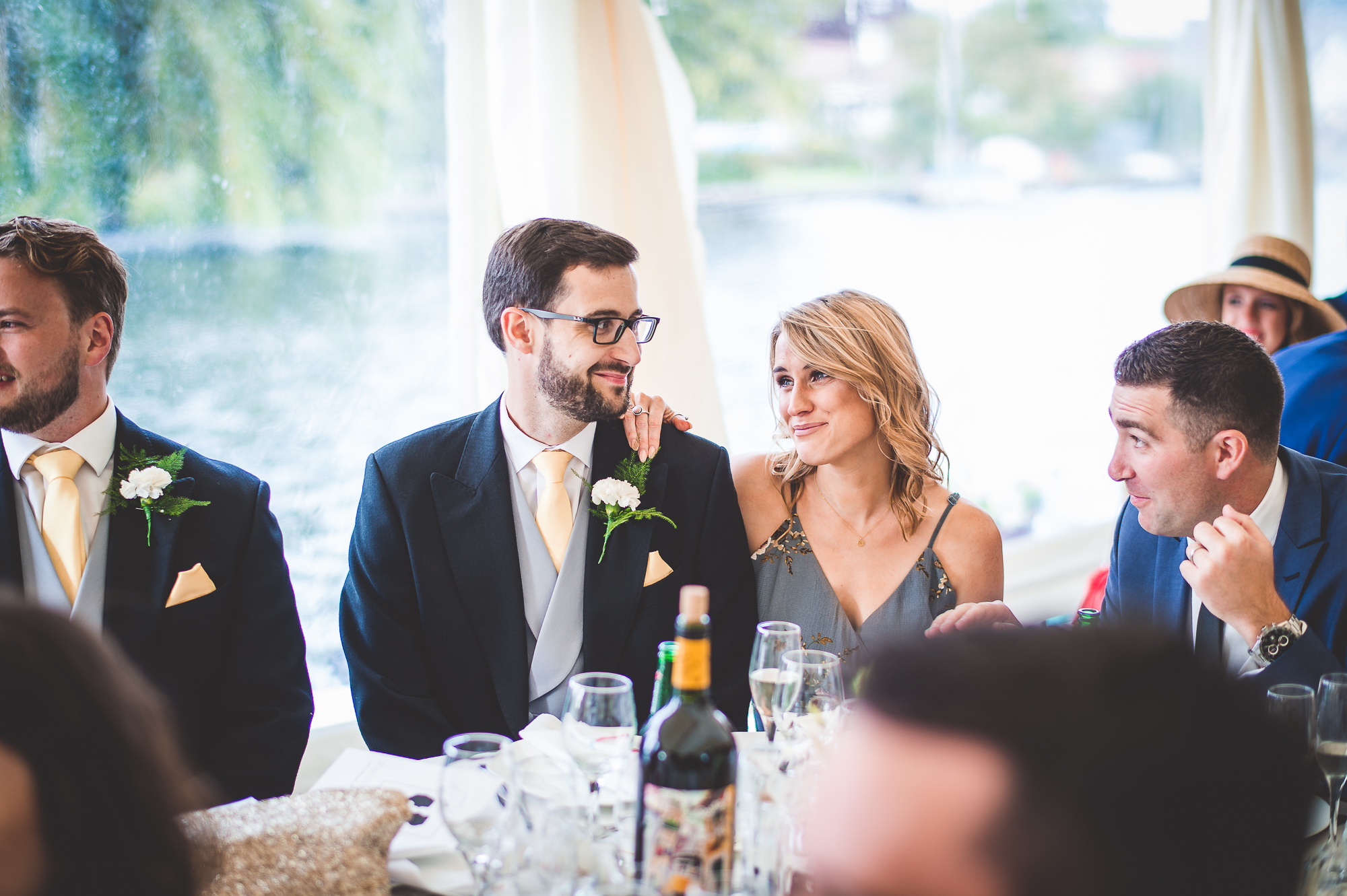 A bride and wedding photographer capture moments of a group sitting around a table at a wedding.