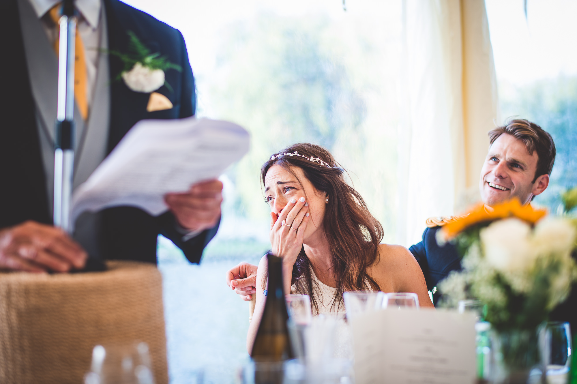 A wedding photographer captures a joyful moment of a bride and groom laughing during a wedding speech.