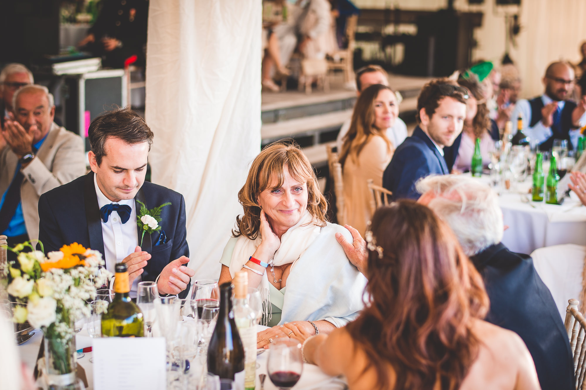 A wedding photo featuring the groom and bride with a group of people sitting at a table.