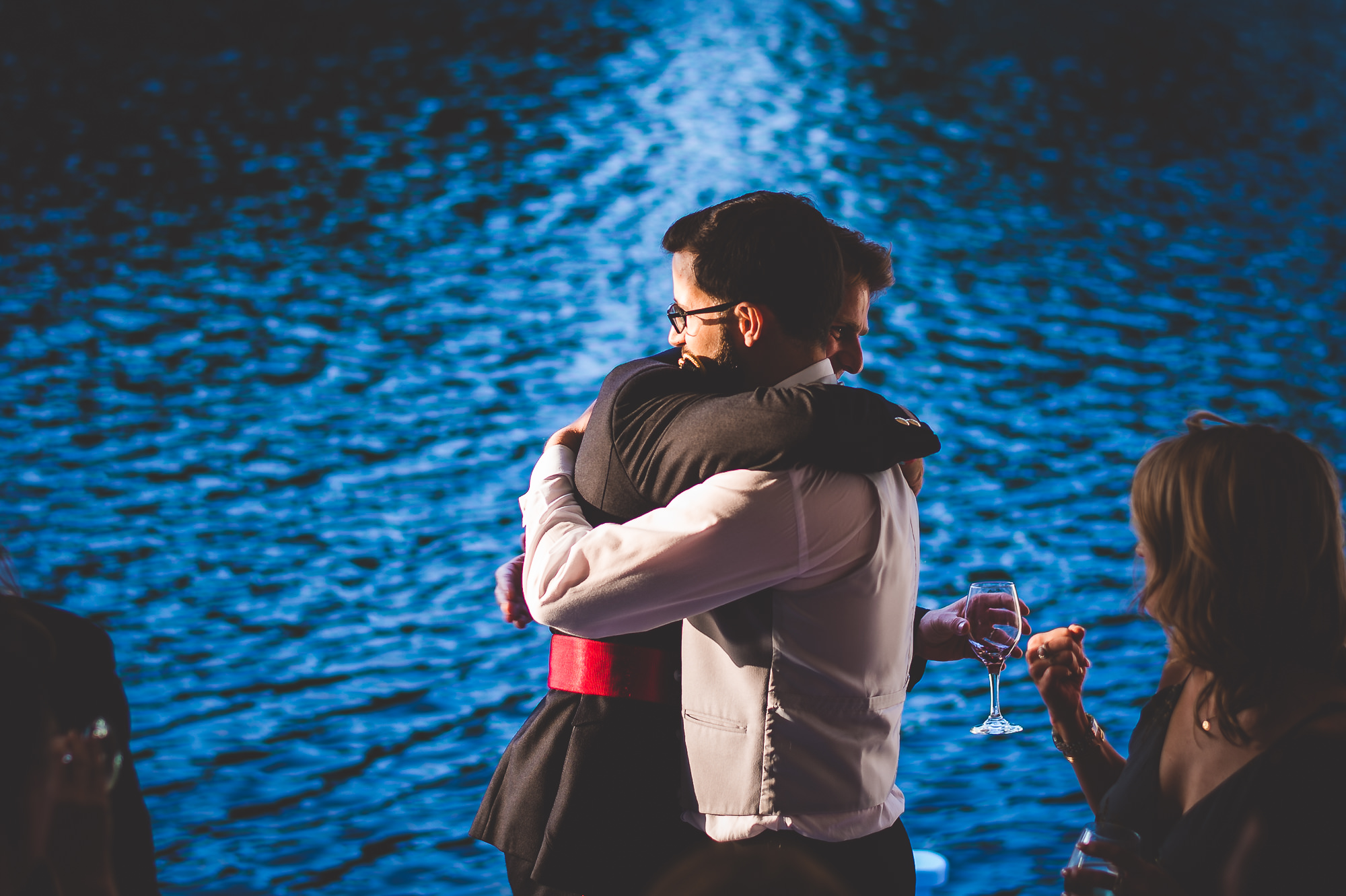 A groom is hugging a woman in front of a body of water, captured by a wedding photographer.