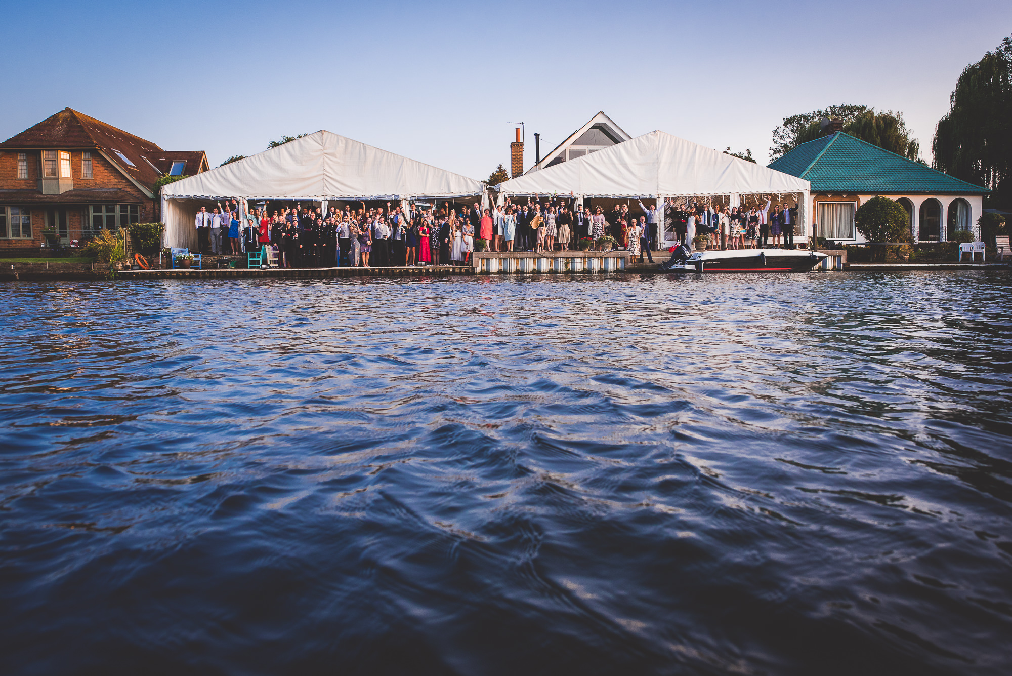 A group of people, including the groom, standing on a boat in front of a house, are captured by the wedding photographer.