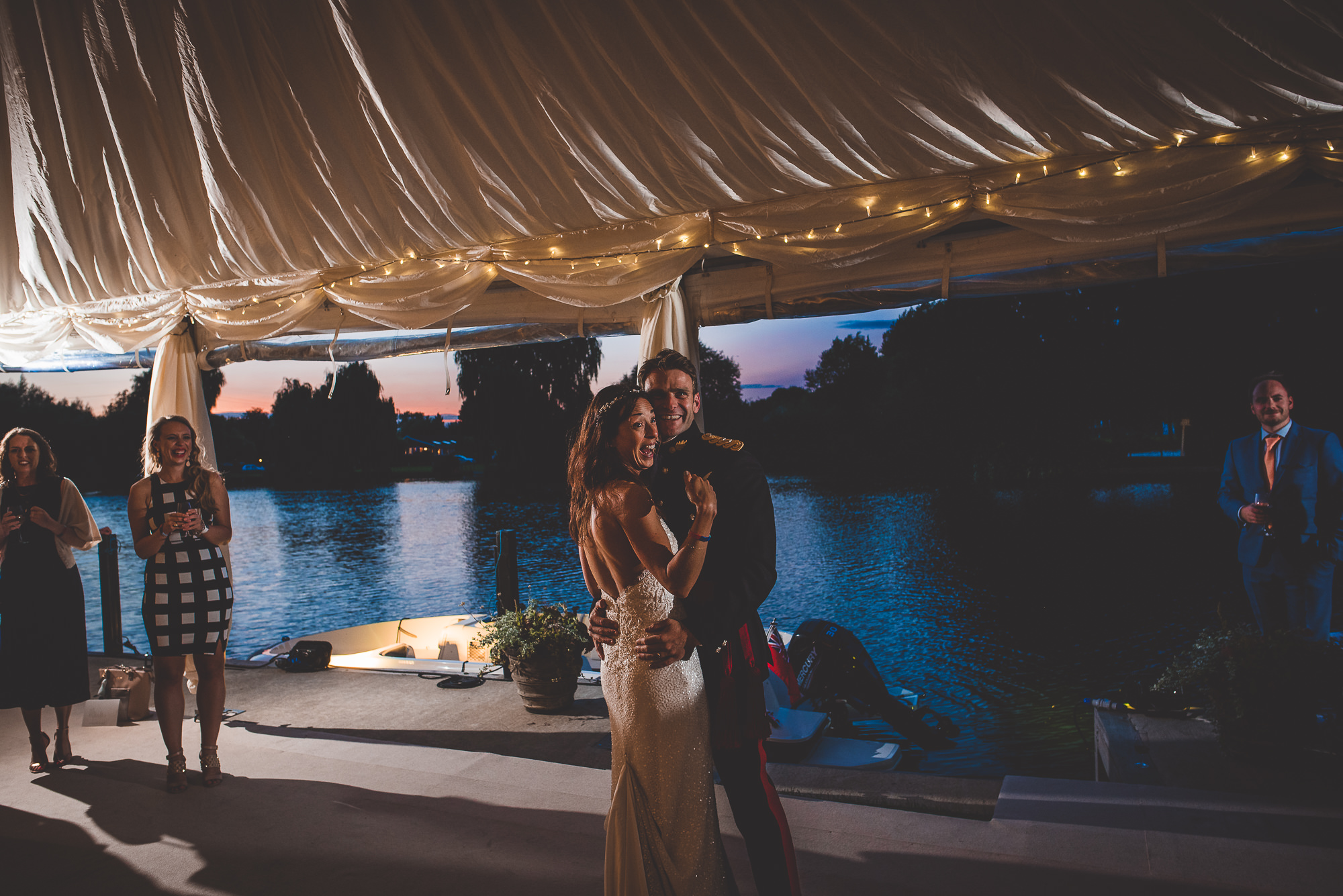 A wedding photo captures a bride and groom gracefully dancing under a tent by a serene lake.