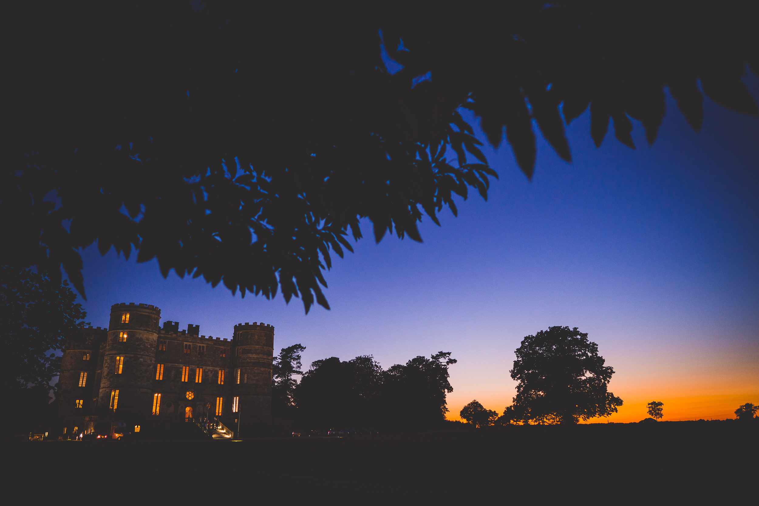 A wedding photographer captures the groom in front of a castle at dusk, with trees in the background.