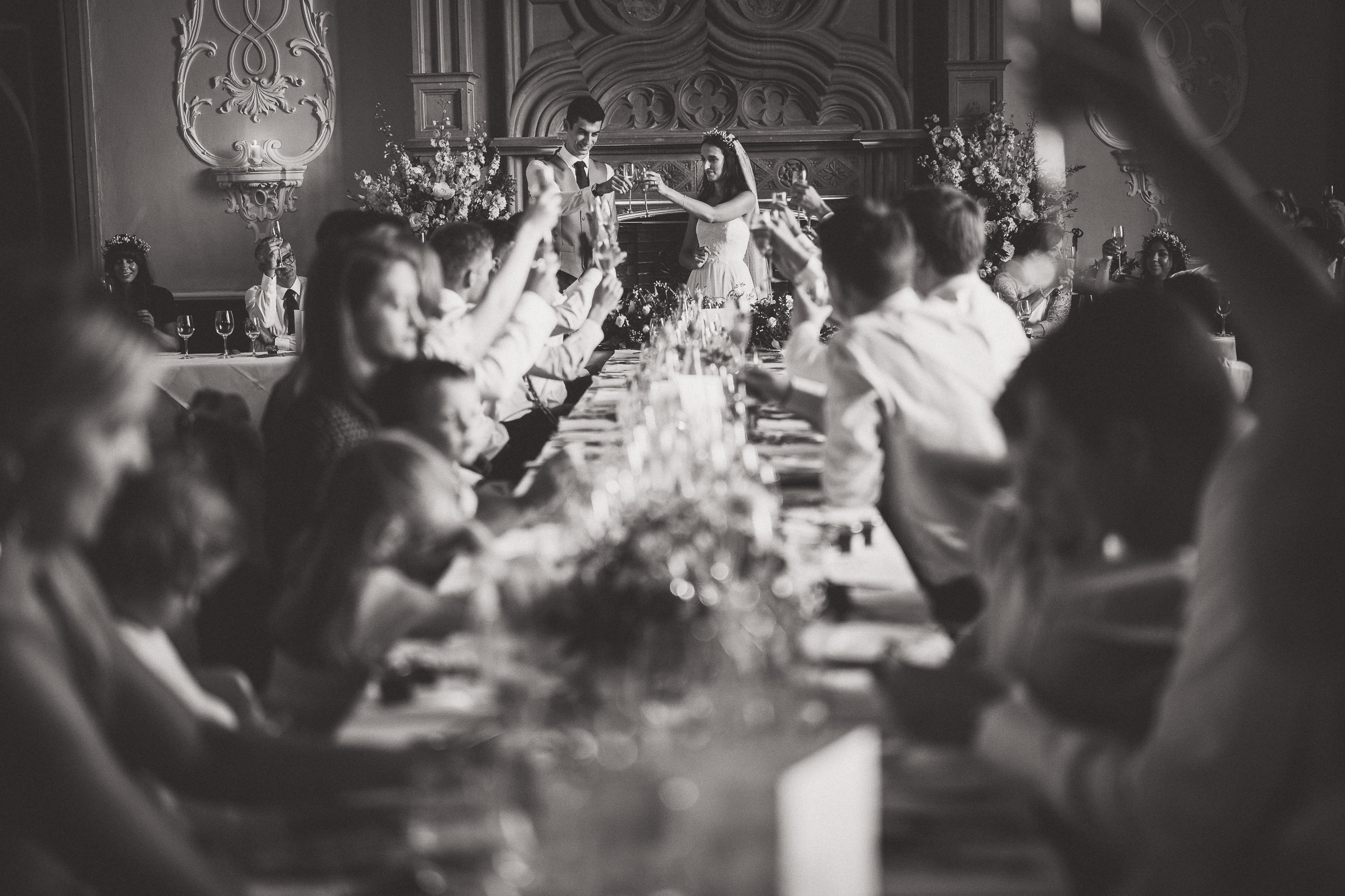 A black and white wedding photo capturing a long table with people raising their hands.