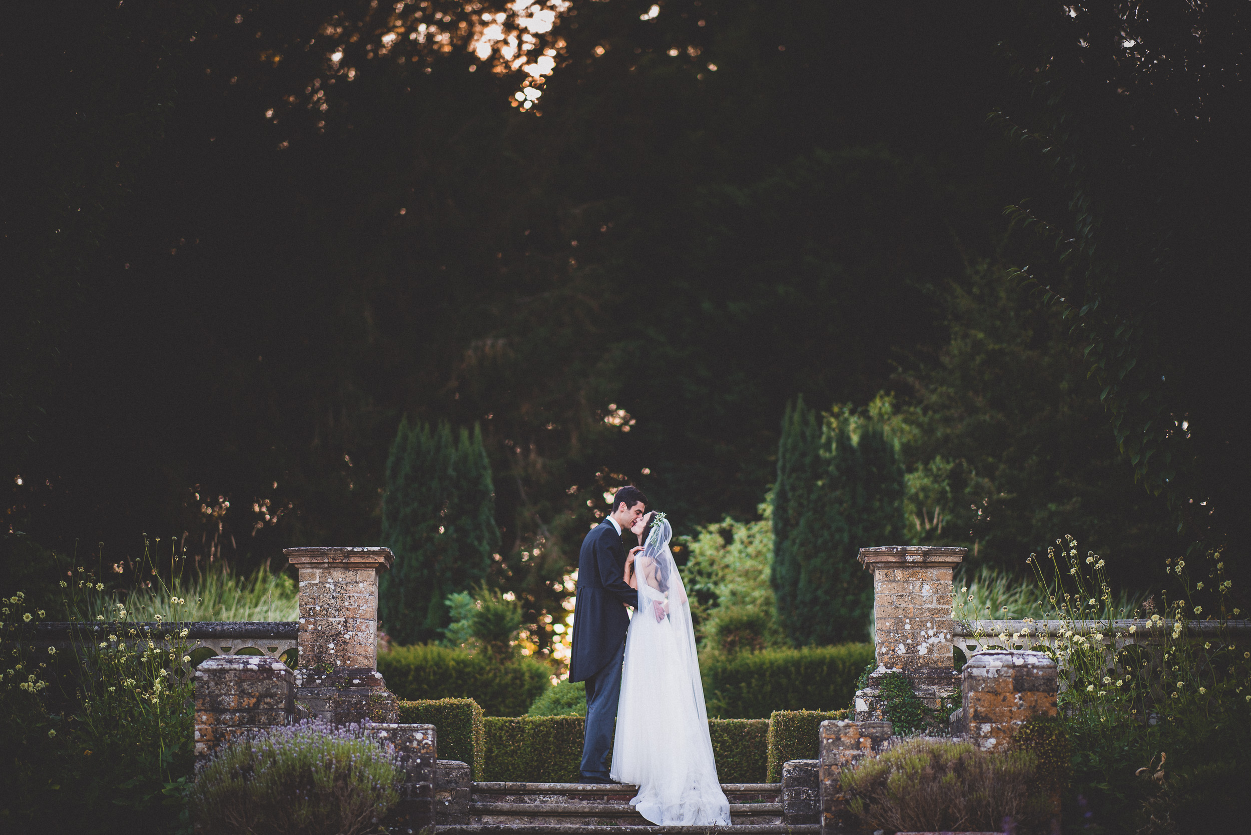 A wedding photographer captures a bride and groom standing on steps in a garden.