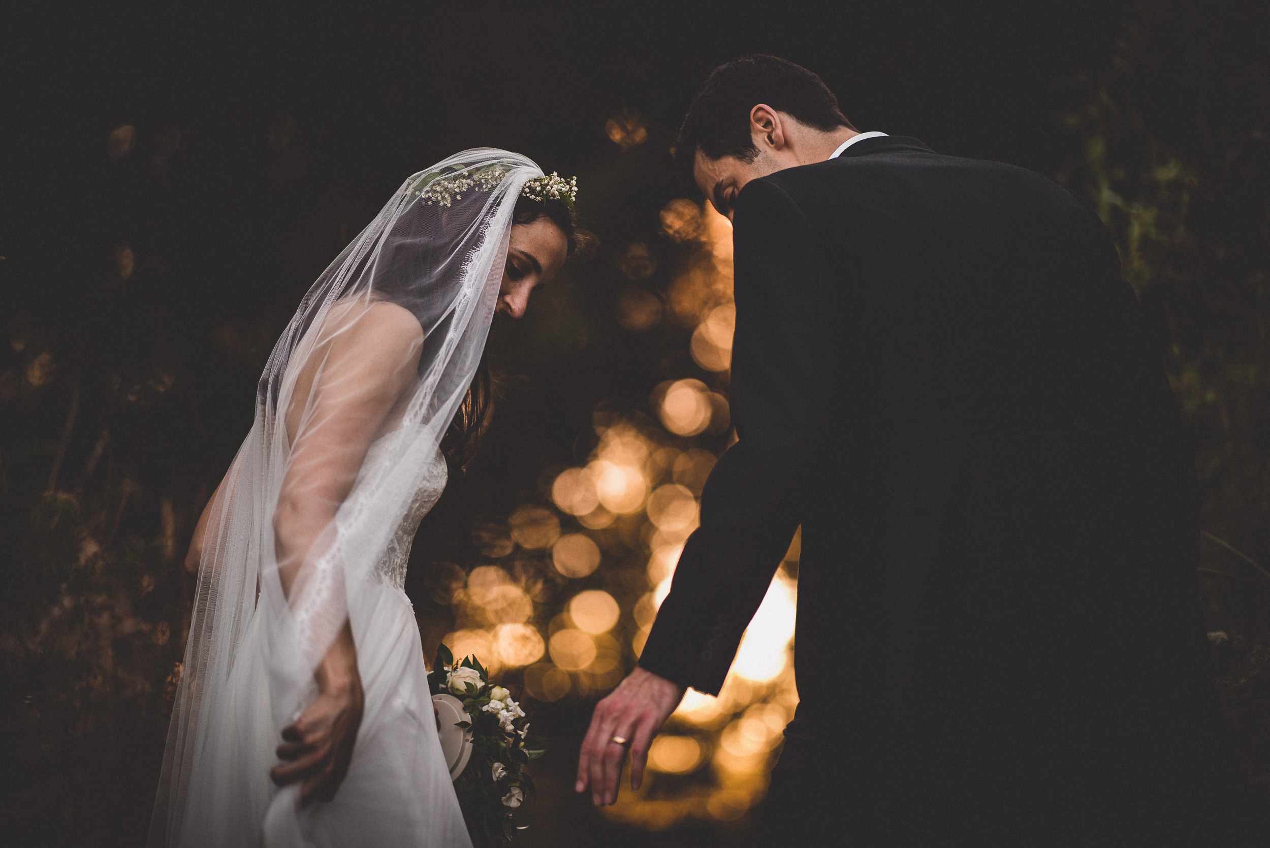 A wedding photographer capturing a bride and groom holding hands in the woods at dusk.