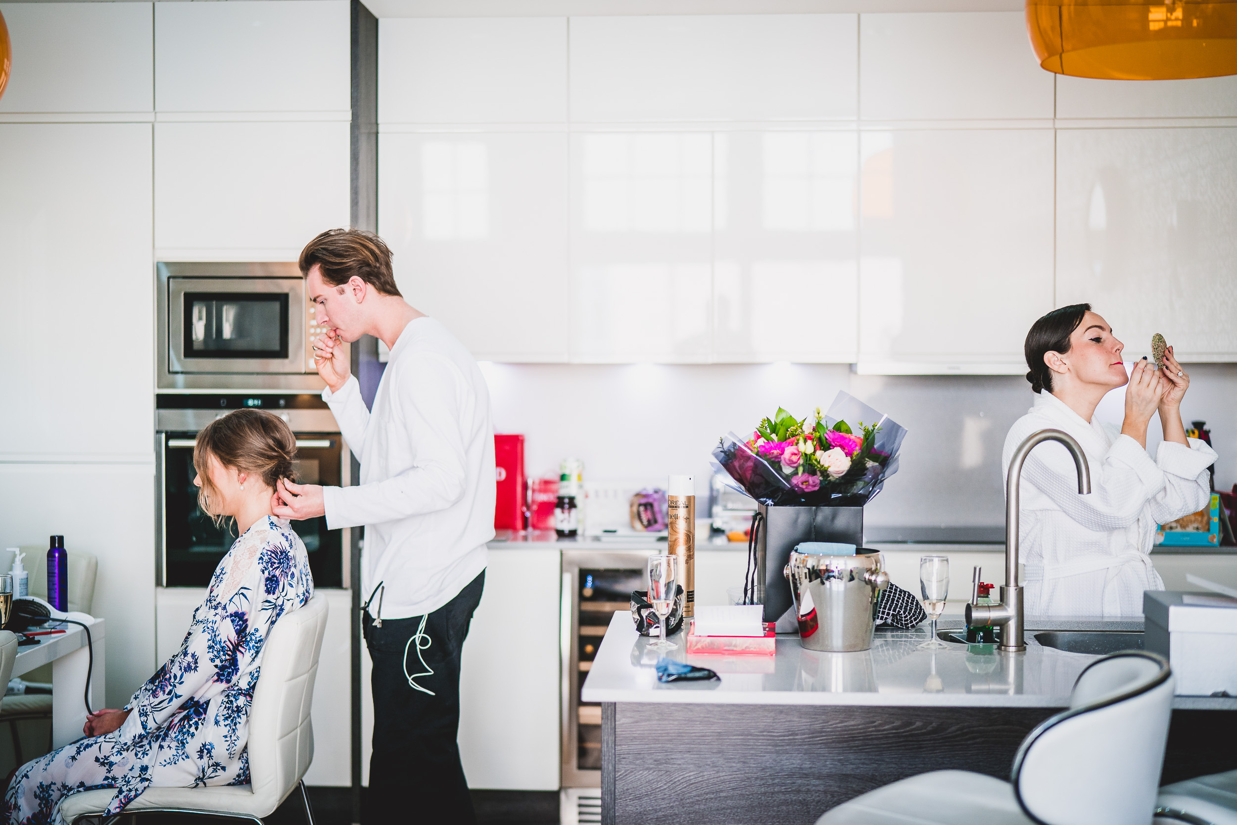 A bride-to-be is getting her hair done in a kitchen, assisted by a wedding photographer capturing beautiful moments for their wedding album.