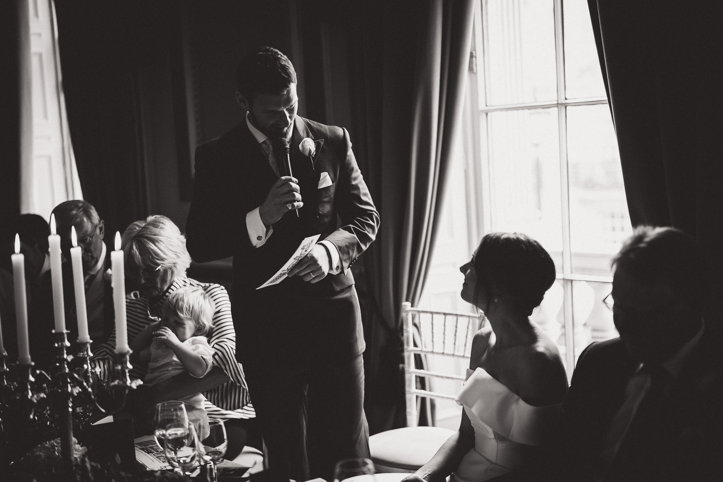 A black and white photo capturing the groom delivering a speech at a wedding.