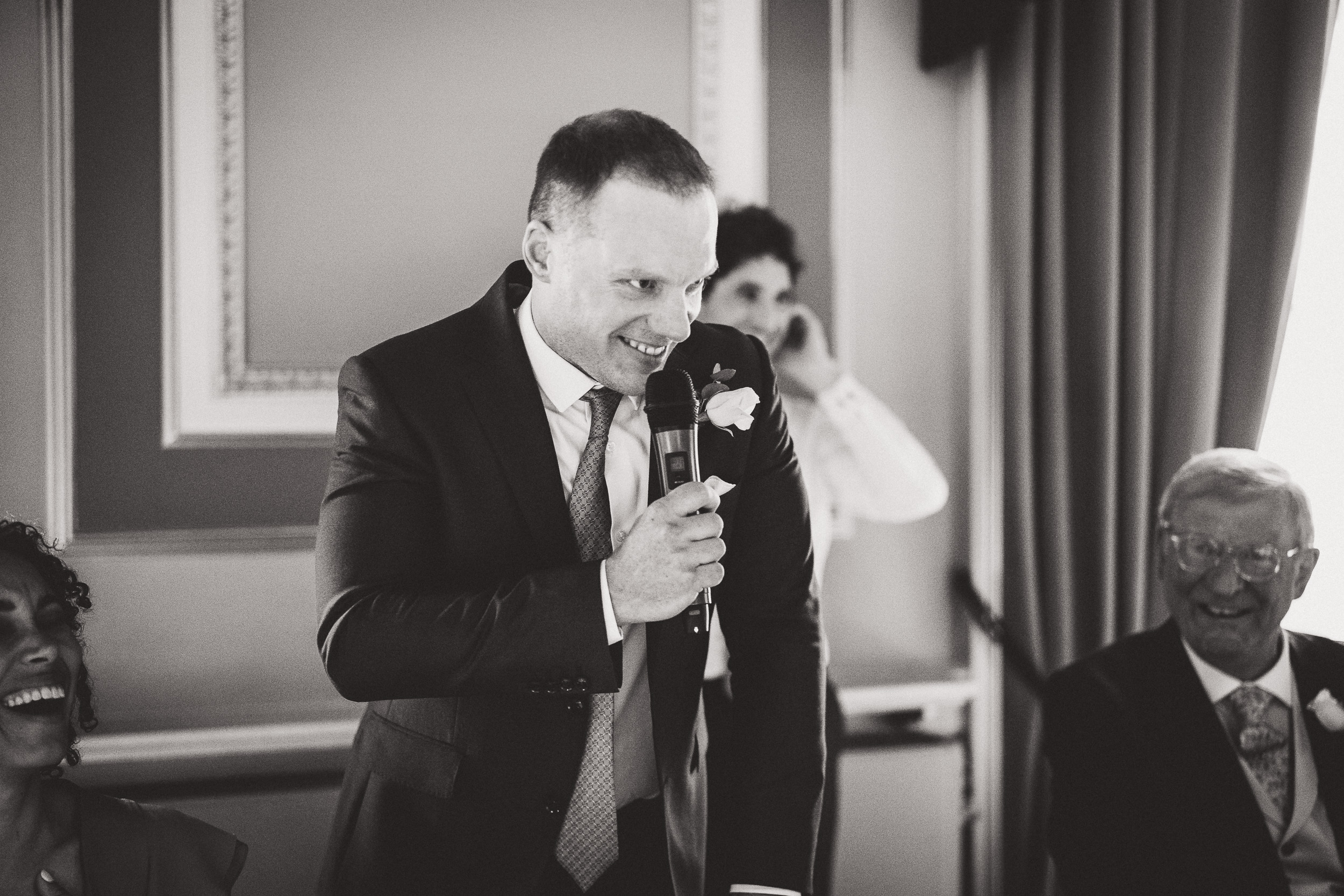 A black and white photo of a man speaking into a microphone at a wedding.