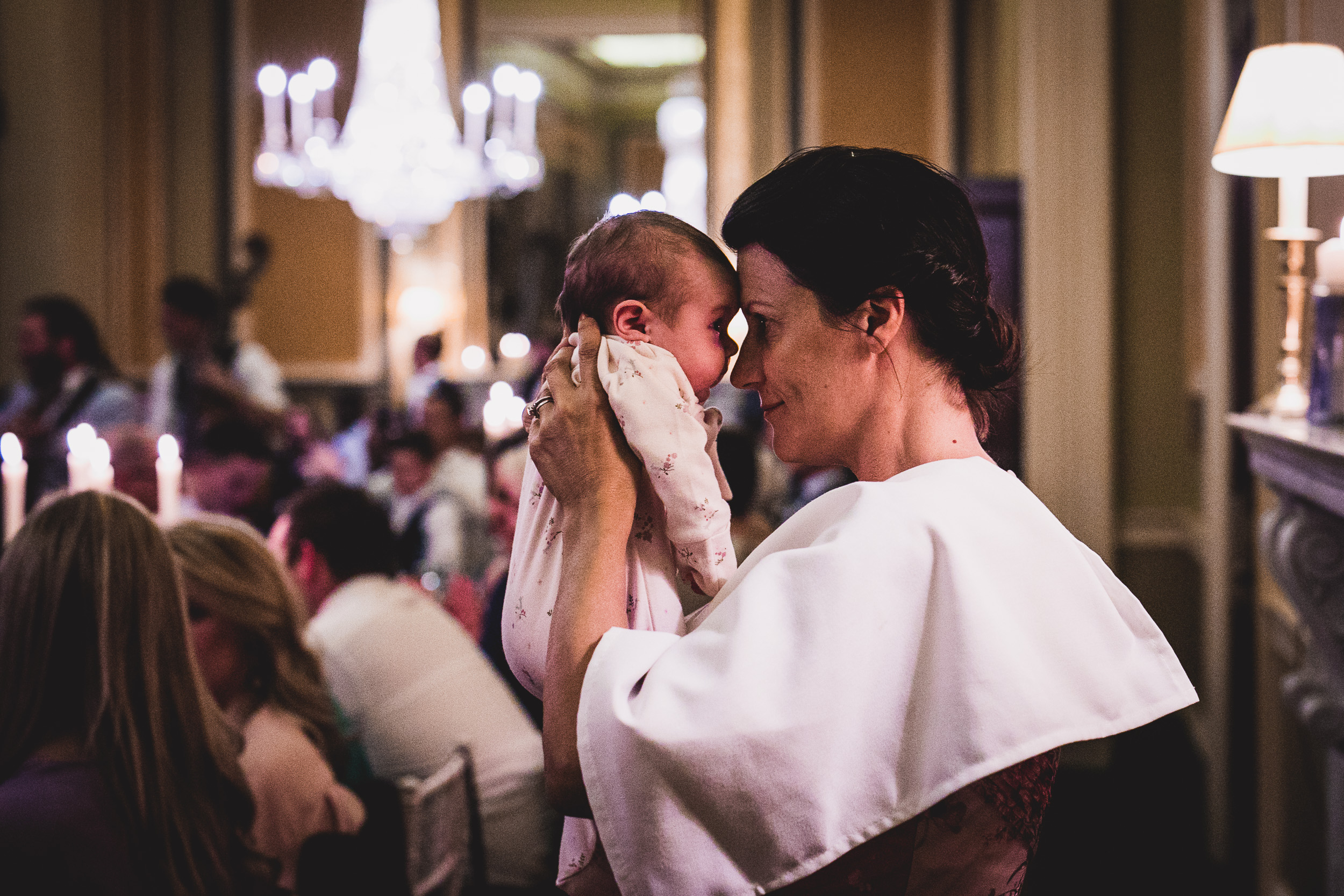 A wedding photographer captures a woman holding a baby in the midst of a crowded room.