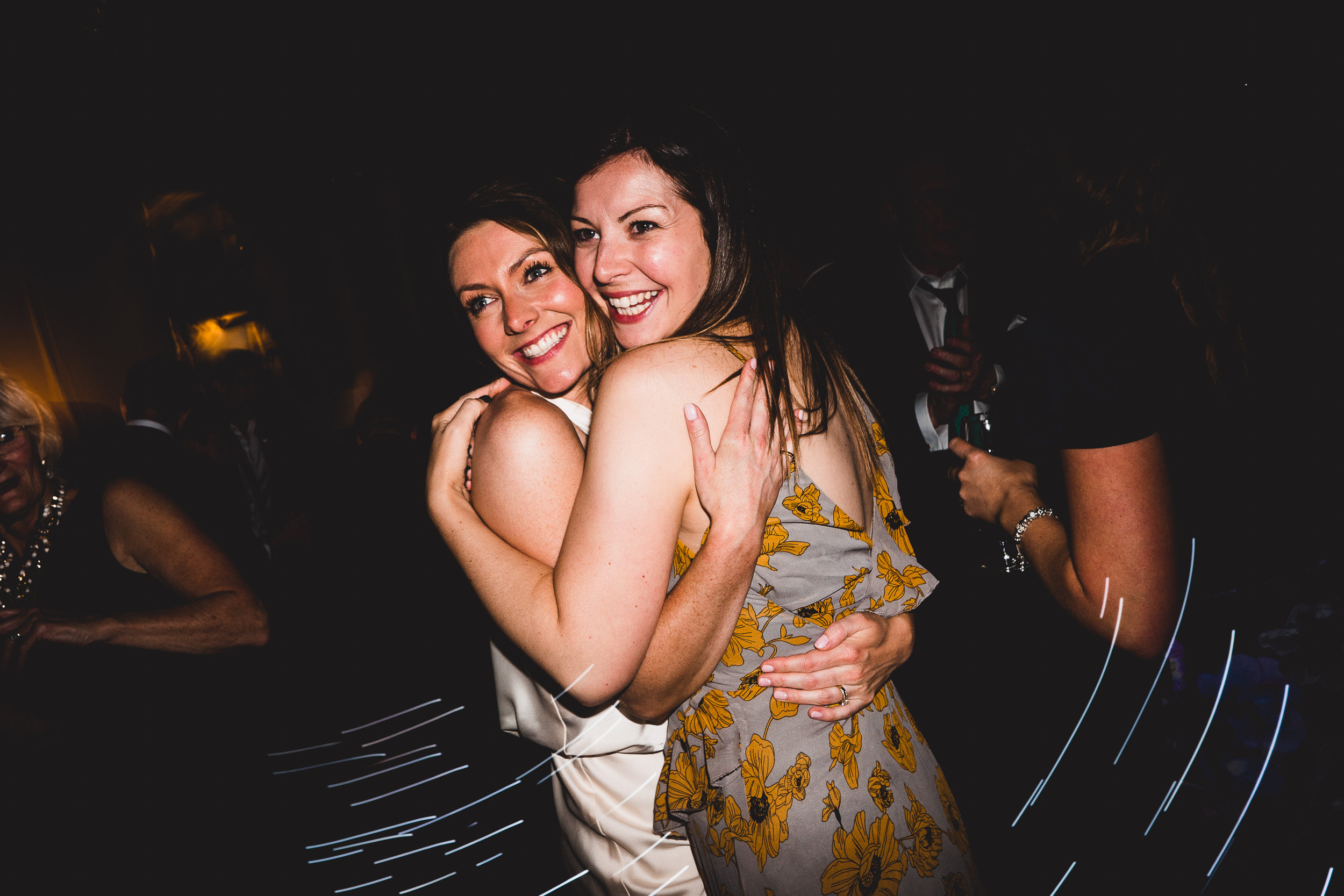 The bride and a friend are hugging each other at the wedding party, with the wedding photographer capturing the heartfelt moment.