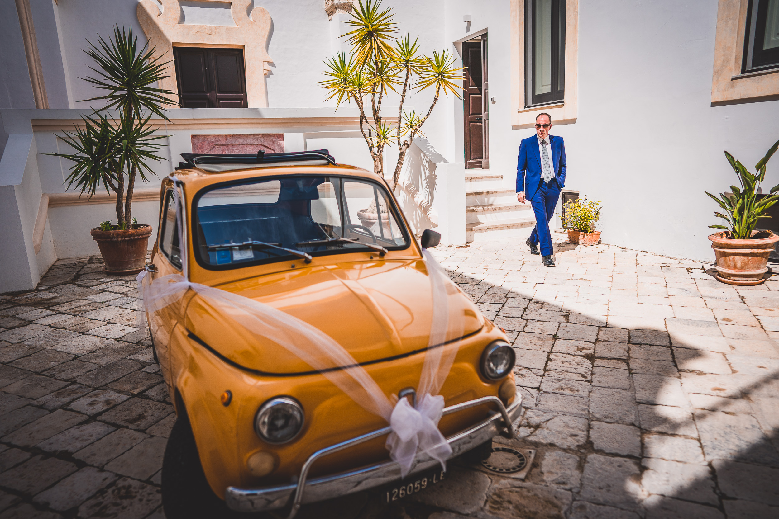 A groom in a suit standing next to a yellow car.