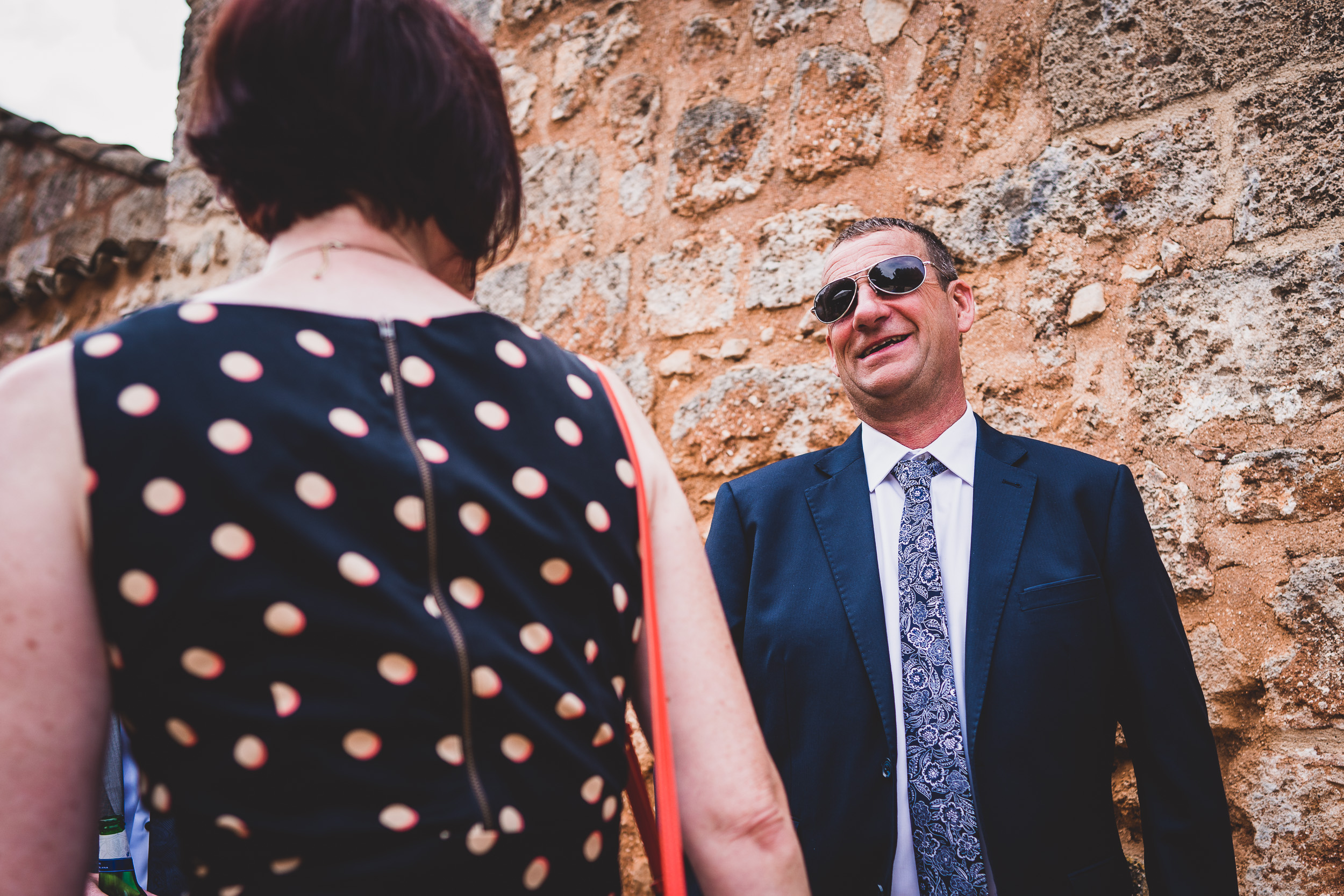 A groom in a suit and sunglasses is talking to a wedding photographer.